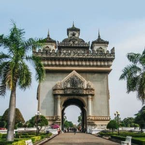 Un arco trionfale in pietra con intagli intricati si erge su un sentiero lastricato, circondato da giardini con palme e panchine bianche.