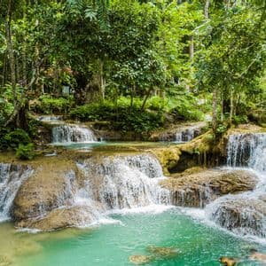 Cascate a gradoni si gettano sulle rocce in una piscina naturale turchese, circondata da una fitta foresta verde, con persone che nuotano.