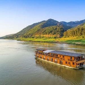 Una barca di legno a due piani naviga su un ampio fiume, affiancata da rigogliose colline verdi sotto un cielo azzurro chiaro.