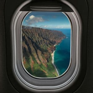 Veduta aerea dal finestrino di un aereo di una costa montuosa e frastagliata con scogliere verdi che si gettano in un oceano turchese.