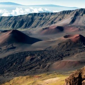 Un paesaggio vulcanico con coni di scorie rossastri e neri sul fondo del cratere, con una catena montuosa sullo sfondo.