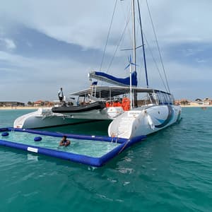 Un grand catamaran blanc ancré dans des eaux turquoise avec une piscine gonflable attachée, où une personne nage près d'une plage de sable.