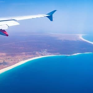 Une aile d'avion vue depuis un hublot, survolant un littoral tropical avec une plage de sable blanc et une mer turquoise.