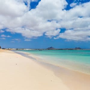 Une large plage de sable blanc rencontre une eau turquoise limpide sous un ciel bleu parsemé de nuages blancs.