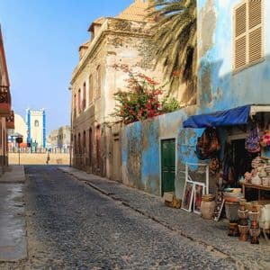 Une rue pavée tranquille bordée de bâtiments colorés et patinés, et un étal de marché vendant de l'artisanat sous un ciel clair.