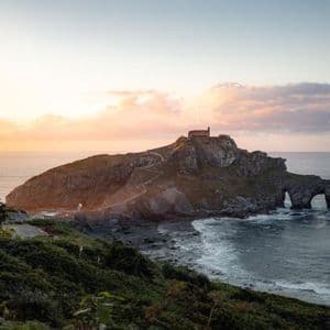 Un isolotto roccioso con un edificio storico in cima, visto da una scogliera verde mentre il sole tramonta sull'oceano.