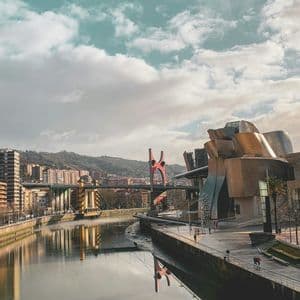 Un moderno edificio architettonico su una riva di fiume con il suo riflesso nell'acqua, un ponte rosso e una città sullo sfondo.