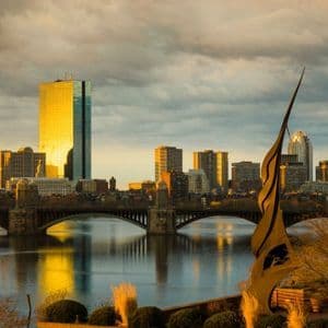Uno skyline cittadino con grattacieli che riflettono la luce dorata del tramonto su un fiume con un ponte.