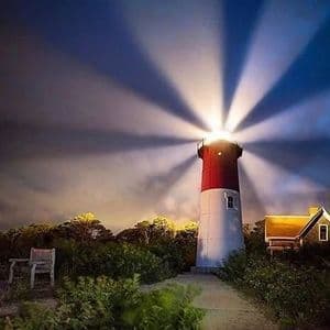Un faro rosso e bianco proietta potenti fasci di luce attraverso un cielo notturno scuro e nuvoloso, con un piccolo cottage e alberi nelle vicinanze.