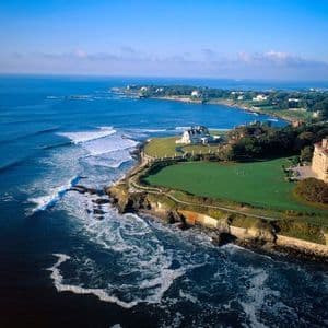 Una vista aerea di una costa rocciosa con grandi ville su prati verdi, mentre le onde si infrangono sulla riva.