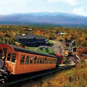 Un trenino funicolare arancione con passeggeri sale su una ripida collina, dominando una vasta foresta con fogliame autunnale e montagne in lontananza.