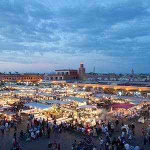 Una vista dall'alto di una vivace piazza del mercato all'aperto al tramonto, affollata di gente e bancarelle di cibo illuminate a festa sotto un cielo nuvoloso.
