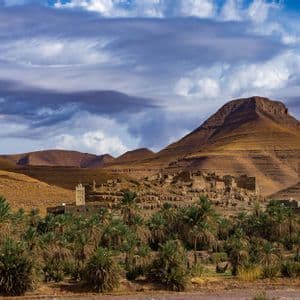 Un antico villaggio e minareto incastonati in una lussureggiante oasi di palme, ai piedi di aride montagne stratificate sotto un cielo nuvoloso.