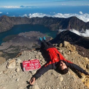 Ein Mann in einer roten Jacke ruht auf einem felsigen Berggipfel und blickt auf einen riesigen Vulkankrater mit einem See darin.
