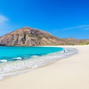 Ein weißer Sandstrand grenzt an eine Bucht mit türkisfarbenem Wasser, im Hintergrund ein großer, karger Hügel unter strahlend blauem Himmel.