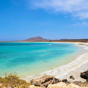 L'eau turquoise de l'océan lèche une plage de sable blanc avec des montagnes arides lointaines sous un ciel bleu clair.
