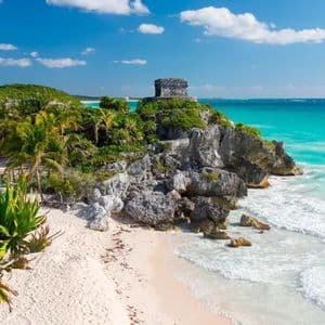 Stone ruins sit on a lush, rocky cliff overlooking a tropical white sand beach and turquoise ocean.