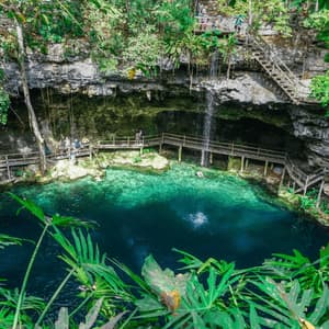 A high-angle view of a WeRoad group trip at a cenote with turquoise water, a waterfall, and wooden walkways built into the rock walls.