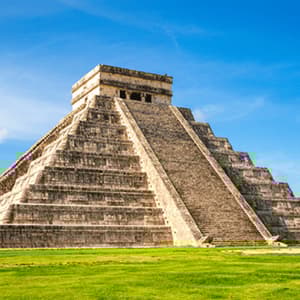A large stone step-pyramid stands on a field of green grass under a bright blue sky with a few clouds.