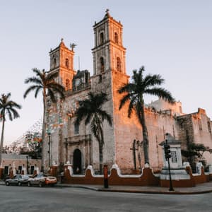 A large stone church with two bell towers glows in the sunset light on a street corner, surrounded by palm trees and passing traffic.
