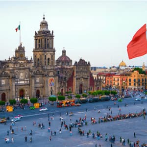A large Mexican flag waves over a bustling historic city square filled with people in front of an ornate cathedral.