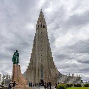 A towering modern church with a stepped concrete facade and a statue on a pedestal in front, under a gray, cloudy sky.