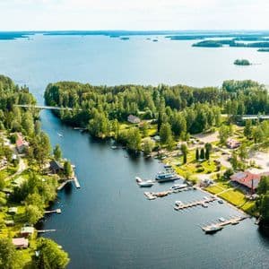 Eine Luftaufnahme eines Wasserwegs, der durch einen dichten Wald fließt, mit einem kleinen Dorf und einem Yachthafen an seinen Ufern, der zu einem großen See mit Inseln führt.