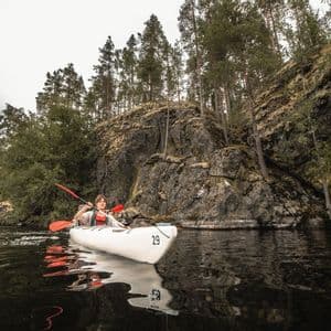Eine Person paddelt in einem weißen Kajak auf dunklem Wasser neben einer felsigen, bewaldeten Küste unter einem bewölkten Himmel.