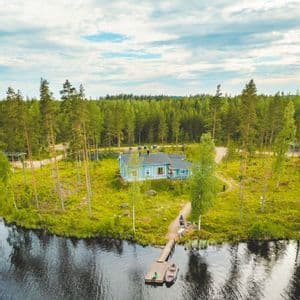 Eine Luftaufnahme eines hellblauen Häuschens auf einer grasbewachsenen Halbinsel, umgeben von einem See und einem dichten Kiefernwald.
