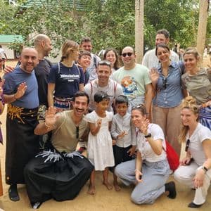 Un gruppo WeRoad in viaggio posa per una foto con due bambini locali, sorridenti e che salutano all'aperto.