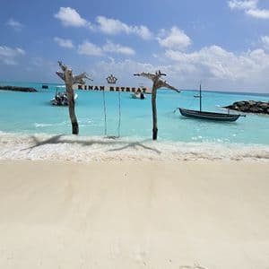 Un'altalena di legno con un cartello 'Kinan Retreat' si erge nell'acqua turchese bassa di una spiaggia di sabbia bianca sotto un cielo blu.