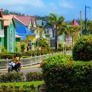 Due persone in scooter percorrono una strada fiancheggiata da case tropicali colorate, palme e siepi fiorite.