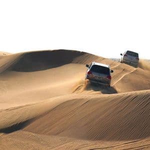 Zwei silberne SUVs fahren über große, rollende Sanddünen in einer Wüste und wirbeln dabei Sand von ihren Reifen auf.