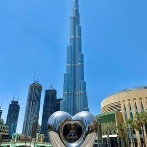 Una grande scultura riflettente a forma di cuore si erge su una piazza con un imponente grattacielo e altri edifici moderni sullo sfondo.