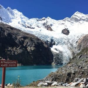 Un cartello di legno per Arhuaycocha accanto a un lago glaciale turchese ai piedi di imponenti montagne innevate e un ghiacciaio.
