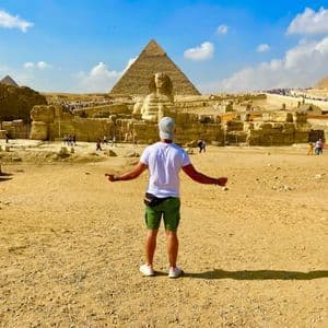 A man seen from behind stands with his arms outstretched, facing the Great Sphinx and pyramids in a desert landscape.
