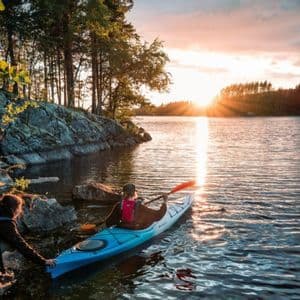 Eine Person an einem felsigen Ufer hilft einer anderen Person in einem blauen Kajak, bei Sonnenuntergang in einen See zu starten.