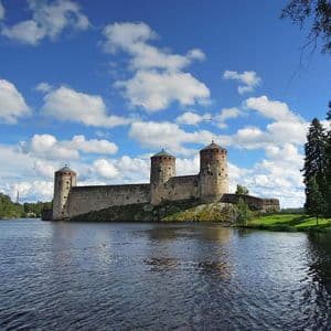 Eine mittelalterliche Steinburg mit mehreren Rundtürmen thront auf einer felsigen Küste neben einem großen Gewässer unter blauem Himmel mit flauschigen Wolken.