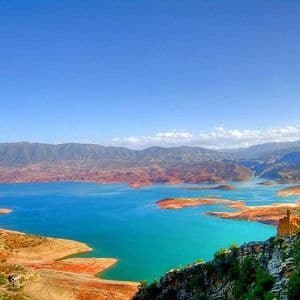 Un lago turchese vibrante si snoda tra colline bruno-rossastre con una catena montuosa sullo sfondo sotto un cielo azzurro limpido.