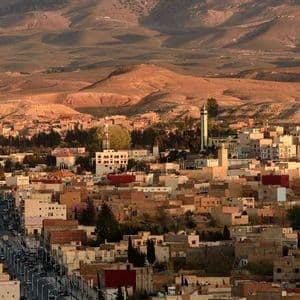 Una vista panoramica di una vasta città con edifici color terra incastonati tra dolci e aride colline al tramonto.