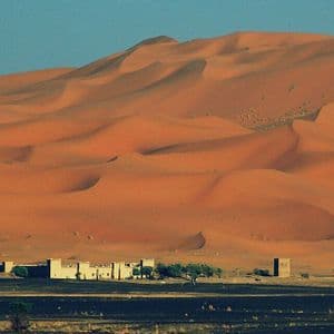 Ai piedi di vaste e ondulate dune di sabbia arancione, sotto un cielo azzurro e limpido, sorge un piccolo villaggio desertico con alberi.