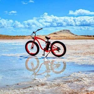 Una e-bike rossa con gomme larghe è parcheggiata in acqua bassa su una salina, creando un chiaro riflesso sotto un cielo blu con nuvole bianche.