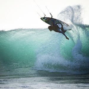Un kitesurfer dai capelli lunghi manovra lungo la cresta di una grande onda oceanica turchese.