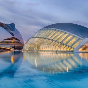 Due edifici moderni e illuminati con architettura curvilinea che si riflettono in un grande e calmo specchio d'acqua al tramonto.