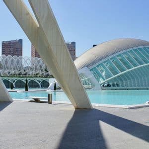 Travi bianche inclinate incorniciano la vista di un grande edificio a cupola futuristico con pareti in vetro, situato accanto a una piscina azzurra brillante.