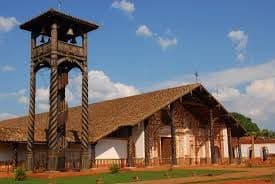 Una chiesa rustica in legno con tetto di tegole sorge accanto a un campanile in legno separato e alto, su un'area erbosa sotto un cielo blu.