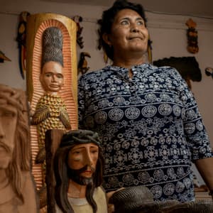 Un'artigiana nel suo laboratorio tra sculture in legno intagliate, come figure religiose e uccelli colorati.