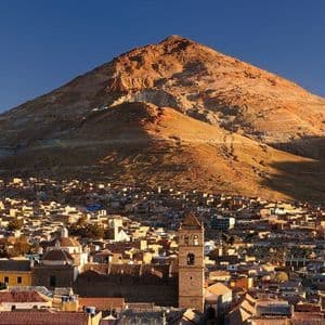 Una città tentacolare con tetti in terracotta e un campanile si trova alla base di una grande montagna illuminata dal sole sotto un cielo azzurro e limpido.