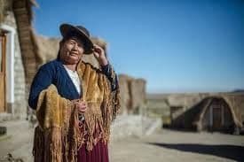 Una donna indigena in abiti tradizionali si erge in un villaggio rurale, toccandosi la tesa del cappello sotto un cielo azzurro e limpido.