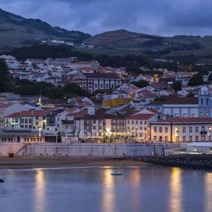 Una città costiera con edifici bianchi e una chiesa blu è illuminata al crepuscolo, con le sue luci che si riflettono sull'acqua.